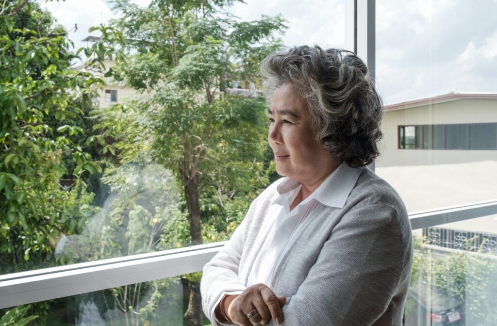 An older adult looks thoughtfully at the green trees outside their window.