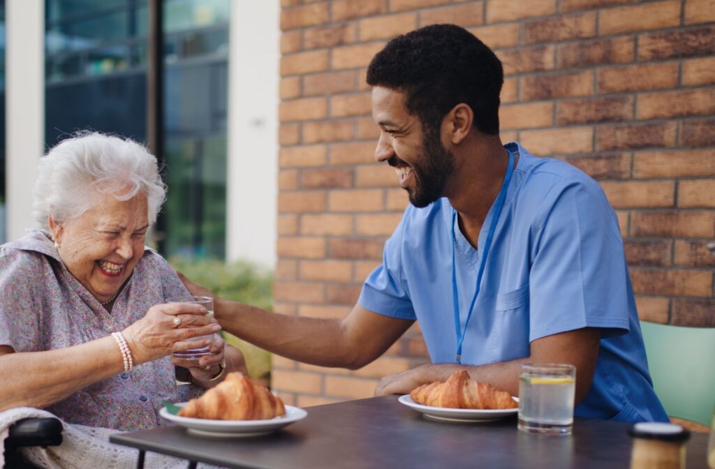 A caregiver helps an elderly woman with breakfast, making a joke so they smile.