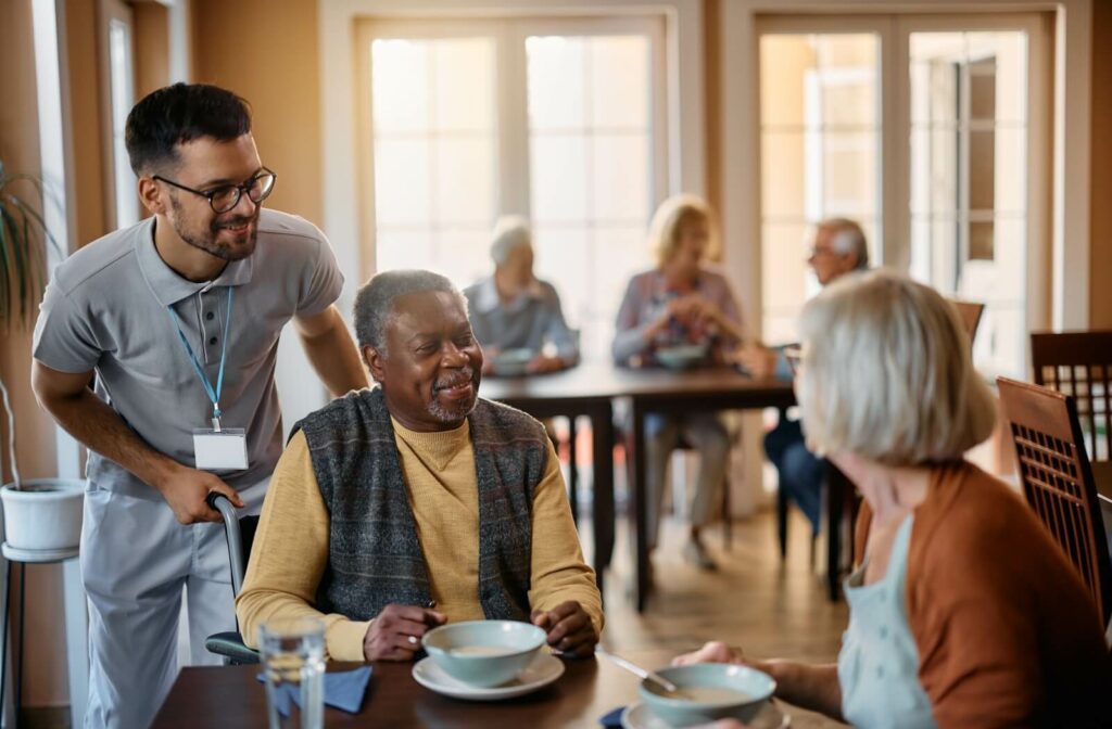 Two seniors dine in their care facility, a caregiver asking them if they would like anything.