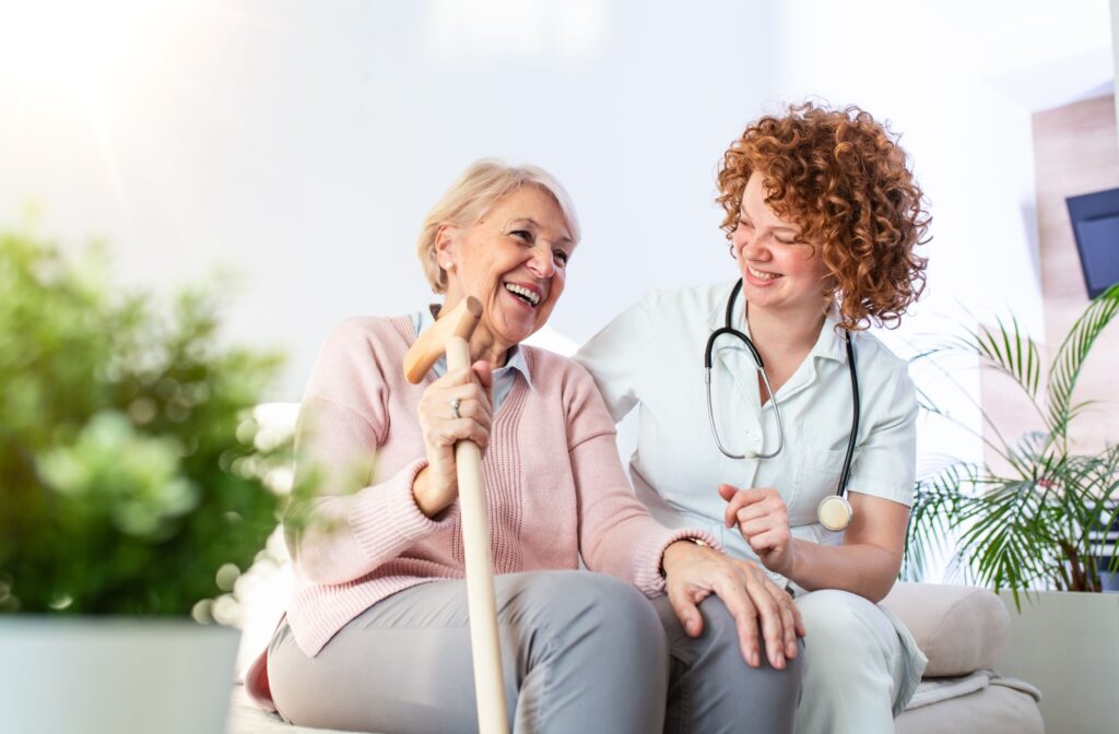 A laughing senior and nurse sit next to each other on a couch in an assisted living community