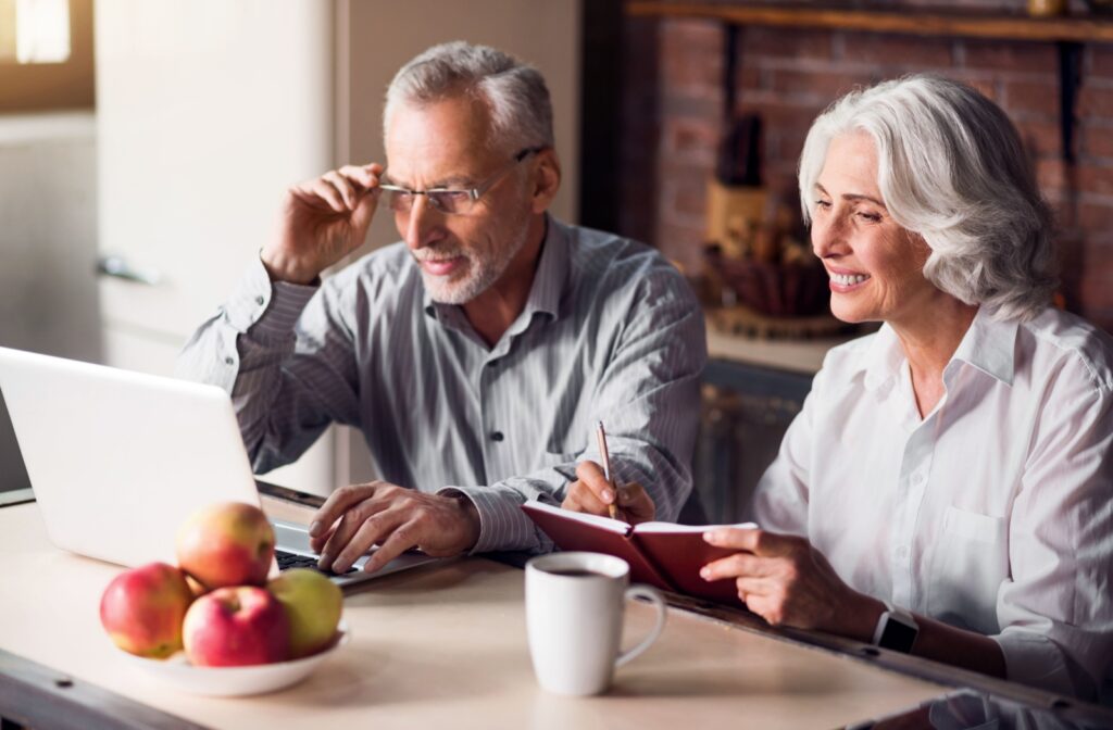 An older adult adjusts their glasses while sitting beside their spouse and researching assisted living communities at their kitchen table