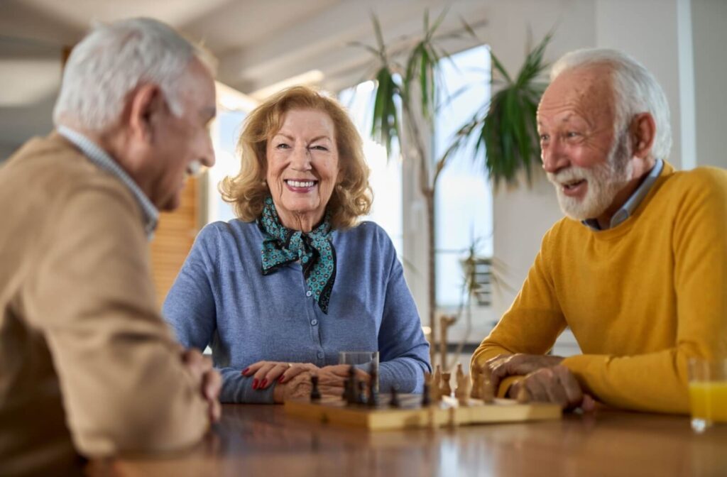 A group of happy seniors talk while playing chess in a senior living community.
