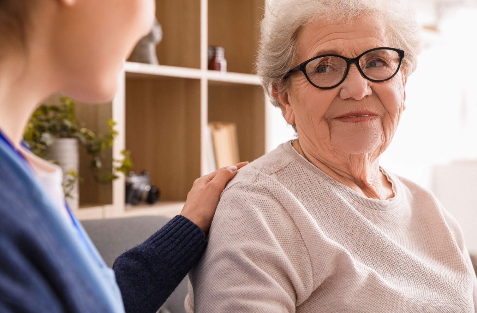 A caregiver in personal care gently touches a smiling older adult's back during a daily check

