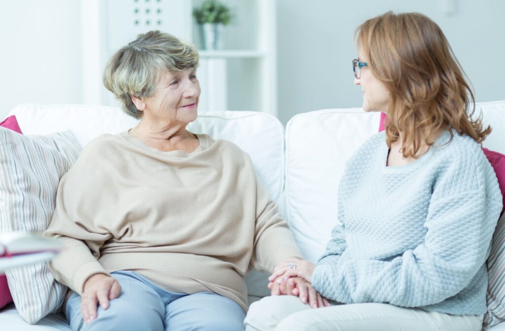 A senior and their younger loved one hold hands as they speak to each other while sitting on a sofa