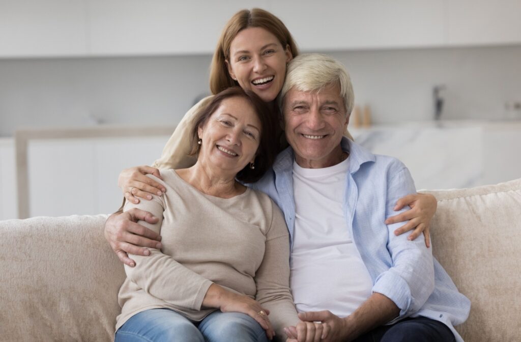 An adult child embraces their smiling senior parents while they sit on a beige sofa